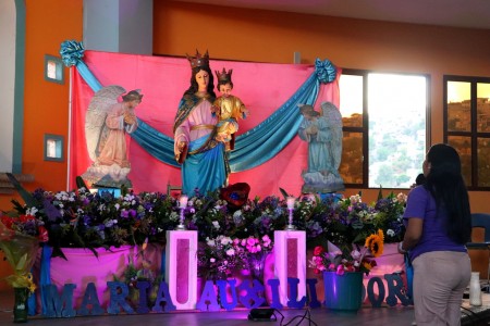 Una joven reza el Santo Rosario con devoción frente al altar adornado de María Auxiliadora, expresión viva de fe y amor filial a la Madre de los jóvenes.